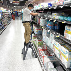 Young man standing on a step stool, stocking shelves of baby products in a grocery story.