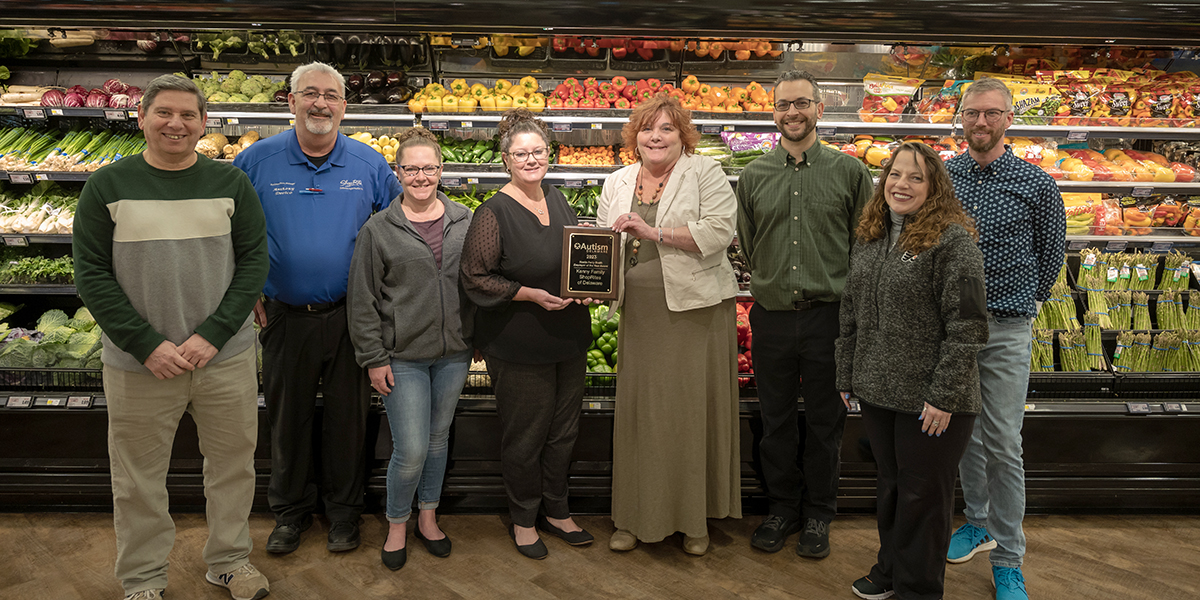 A group of eight smiling people in a grocery store produce section. Two women in the center are holding an award plaque.